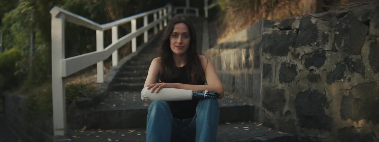 A woman sits on steps outdoors with a stone wall and white railing beside her