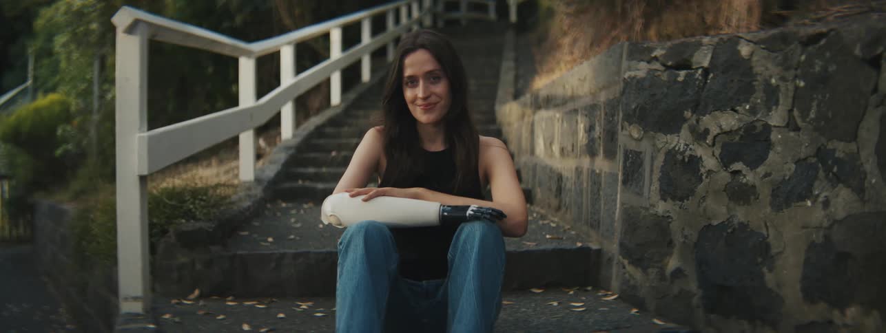 A young woman is smiling while sitting on a step of a staircase and has an arm prosthesis