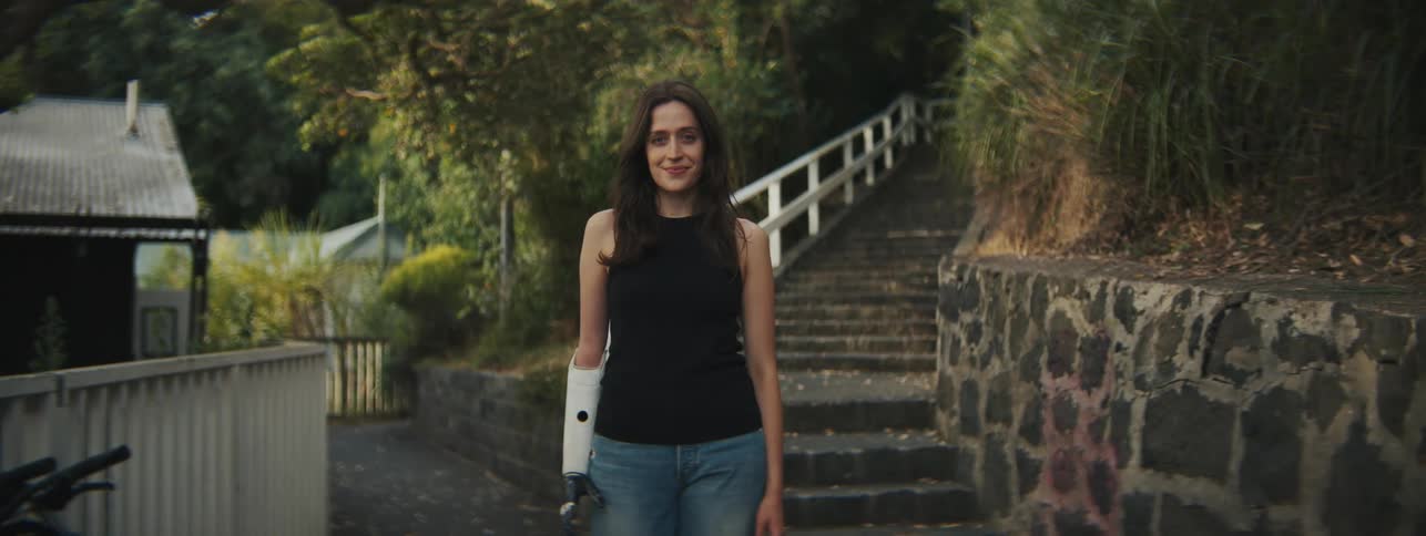 A woman with a prosthetic arm smiles as she stands in front of the stone steps leading to a park
