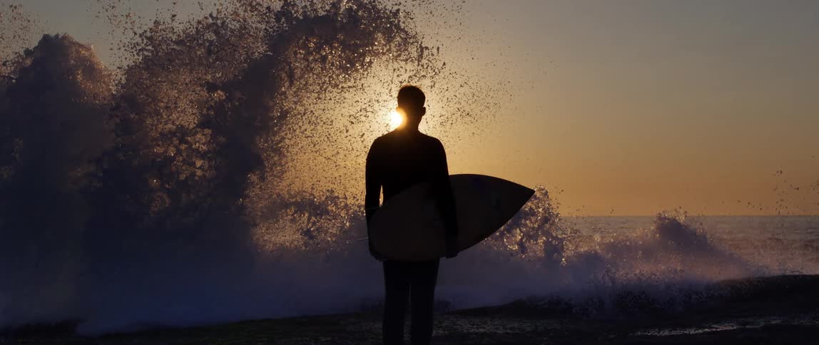Silhouette of a man on the coast with his surfboard and a wave breaking in slow motion in front of him with a sunset on the horizon