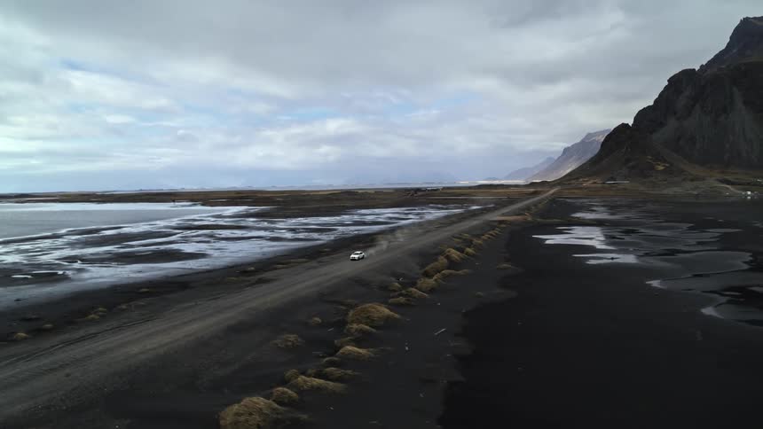 Aerial panorama of a white vehicle speeding along a dirt road on the edge of volcanic mountains and lakes
