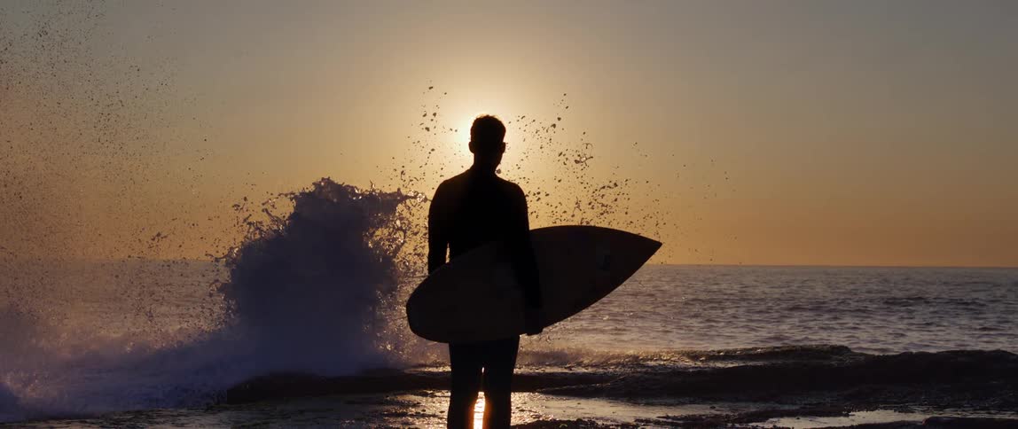 A man carrying a surfboard watches the wave crash