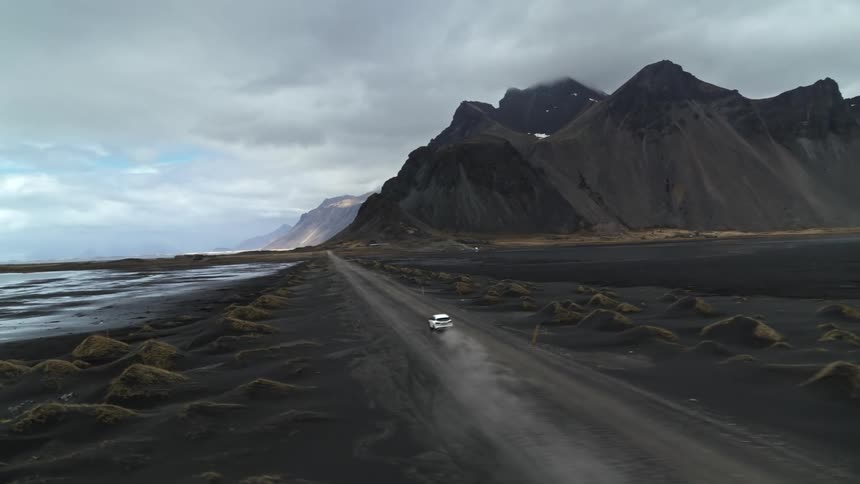 White car speeding along a dirt road next to volcanic mountains on a cloudy day