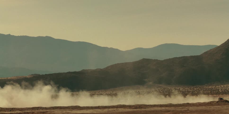 Vehicle driving across the arid desert bordering the mountains kicks up a trail of dust