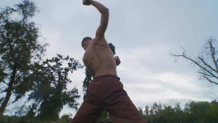 A shirtless man wearing dark pants dancing in a park