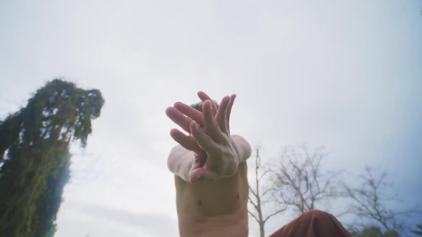 A man dancing using his hands clasping them together and stretching while outside with trees behind him
