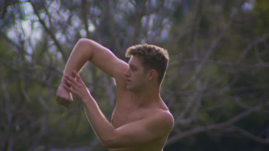 Shirtless man raising up arm and bending back in a dance like motion while standing in a field in front of tree withe tangled bare branches