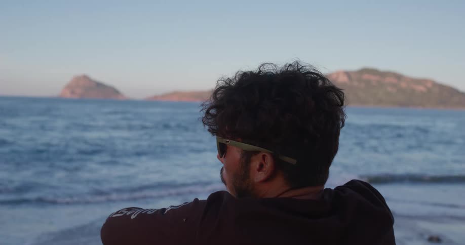A man with sunglasses sits on a coast with mountains in the distance