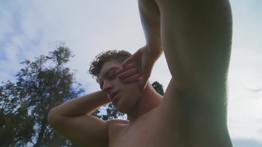 A shirtless male is standing outdoors and stretching his arms