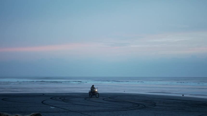 Man riding a motorbike on the wet sand next to the sea in a morning sky
