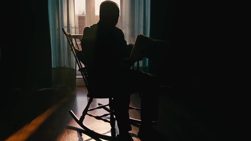 Silhouetted person sits in a rocking chair drinking a coffee reading a a newspaper