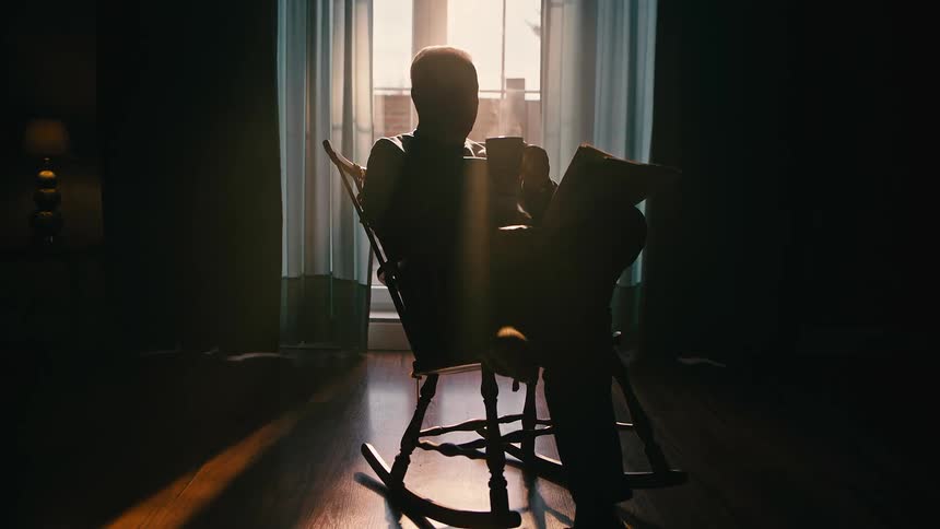 An elderly man sits in a rocking chair beyond a pair of curtains drinking a hot drink and reading a newspaper then gets up