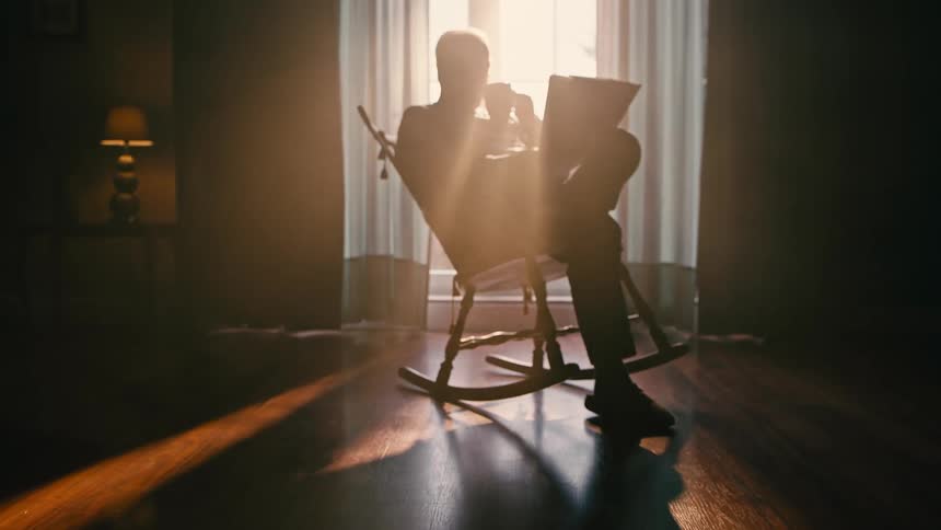 A man sips a drink while sitting in a rocking chair reading a paper