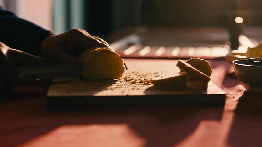 Hands of a person with a knife cutting a slice of bread on a wooden board on the table
