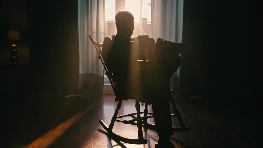 Person silhouetted against a window in a rocking chair reading a newspaper