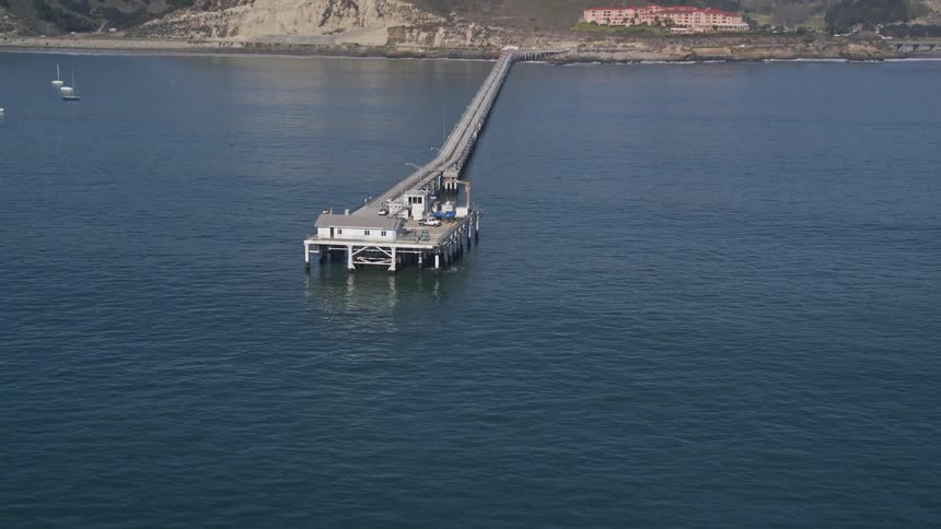 Aerial view of a long pier at sea with yachts around and mountains on the horizon