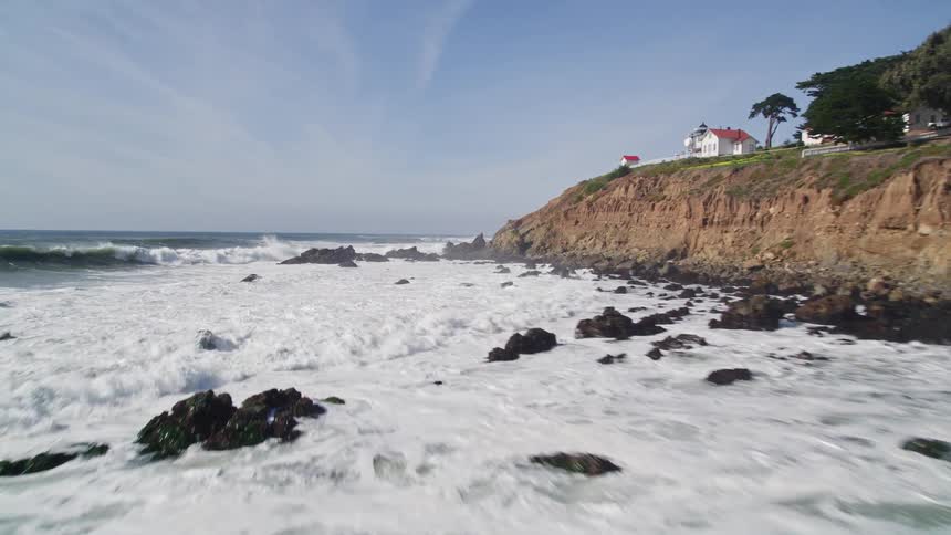 Waves roll into shore and splash against rocks as houses along the coast overlook the water