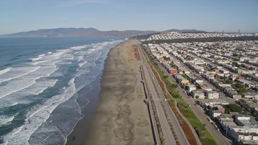 Waves roll in at the shore near a city with mountains behind