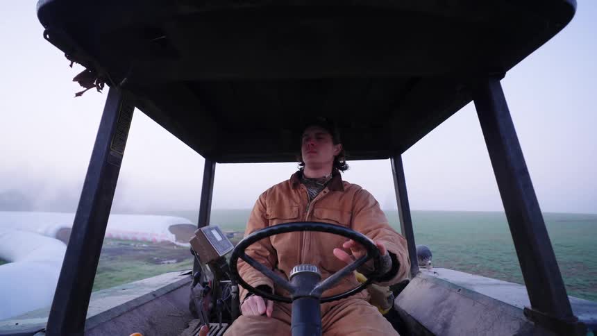 A young farmer drives a tractor on a farm covered in morning mist