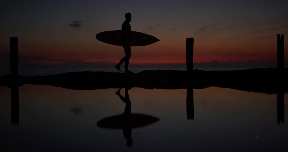 Water reflects surfer silhouetted against the red horizon while carrying a surfboard