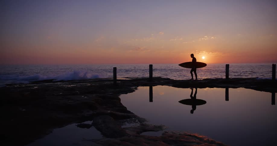 A surfer walking by a tide pool next to a beach at sunset