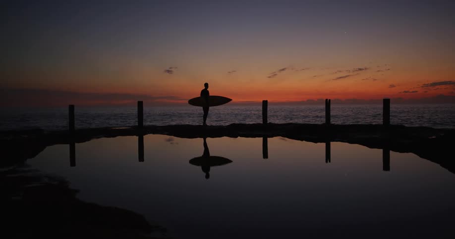 Silhouette of a man walking on a rock by the sea with his surfboard on a morning with an orange sky
