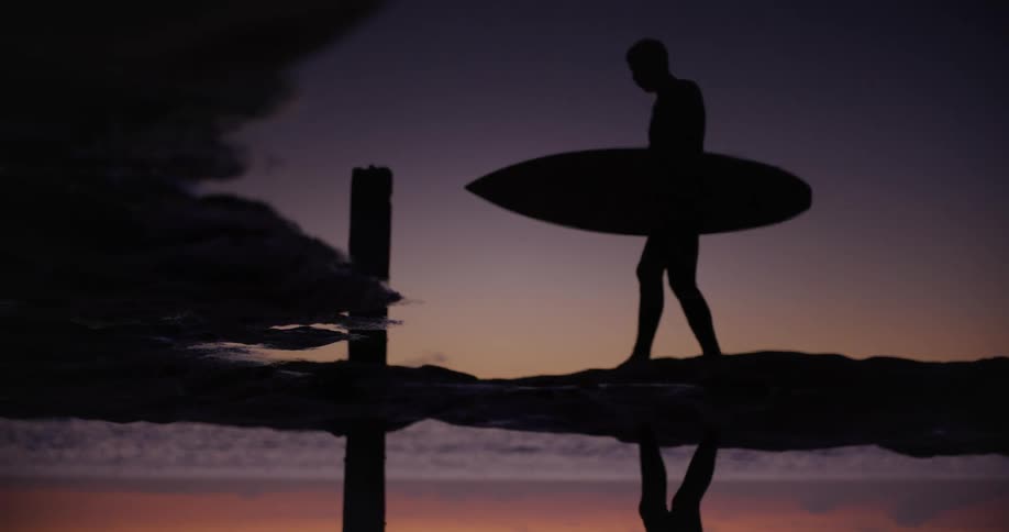 Reflection of the silhouette of a person walking with a surfboard near the coast at sunset