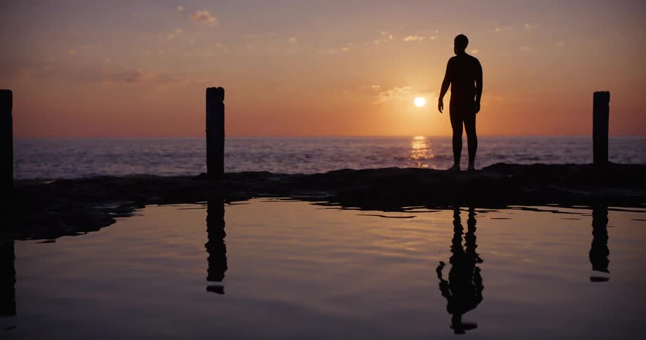 Silhouette of a man standing next to a tidal pool by the beach at sunset