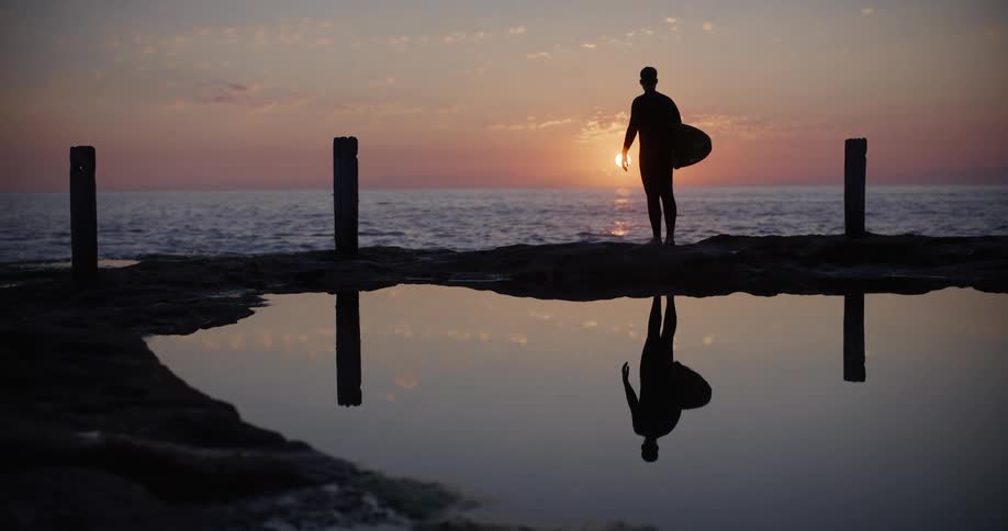 Man on the rock holding his surfboard overlooking the sea with sunrise on the horizon