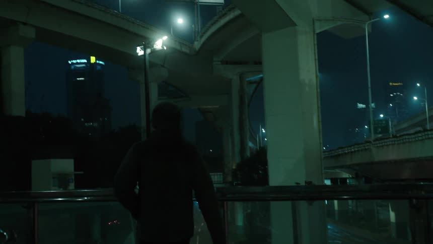 Silhouette of a person wearing a hoodie walking under an overpass in the city at night