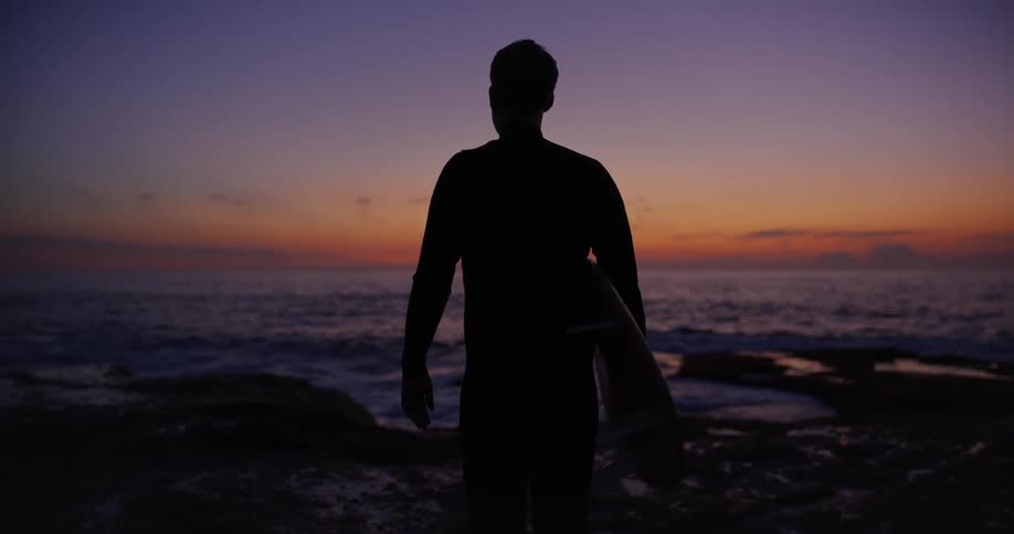 Silhouette of a man from behind holding his surfboard while standing on a rock by the sea in a morning with reddish and purple sky