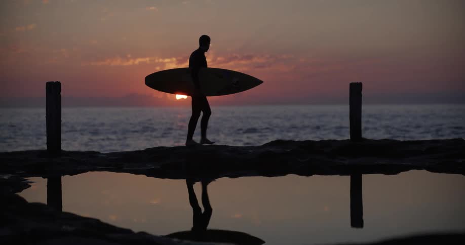 Man carries his surfboard as he walks on the beach and the sun glows on the horizon