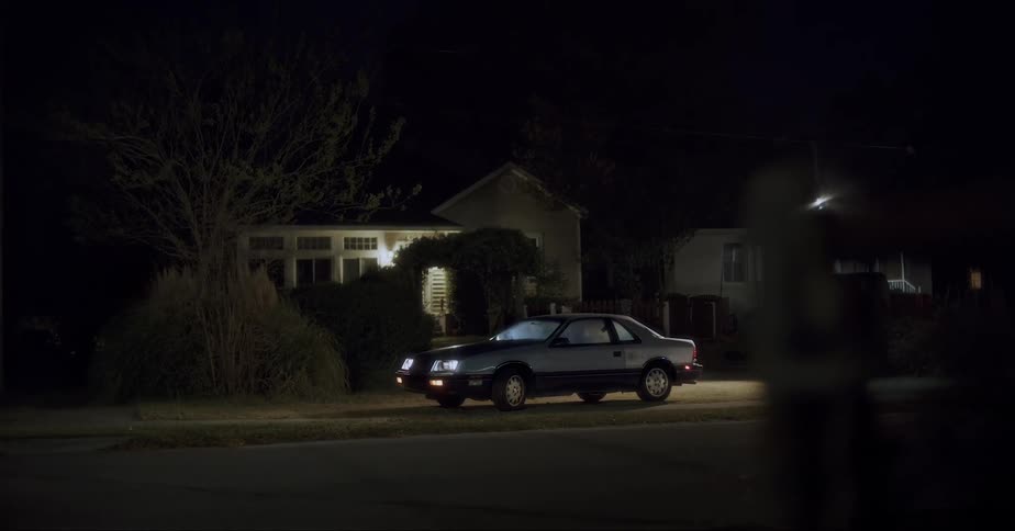 Car with its lights on sits parked on lawn and sidewalk near house at night