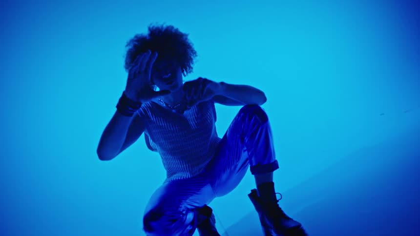 A sexually ambiguous person squats and gestures in a front of a blue screen