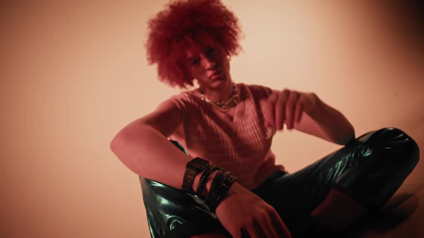 A person with a red afro is sitting wearing a striped shirt and shiny green pants