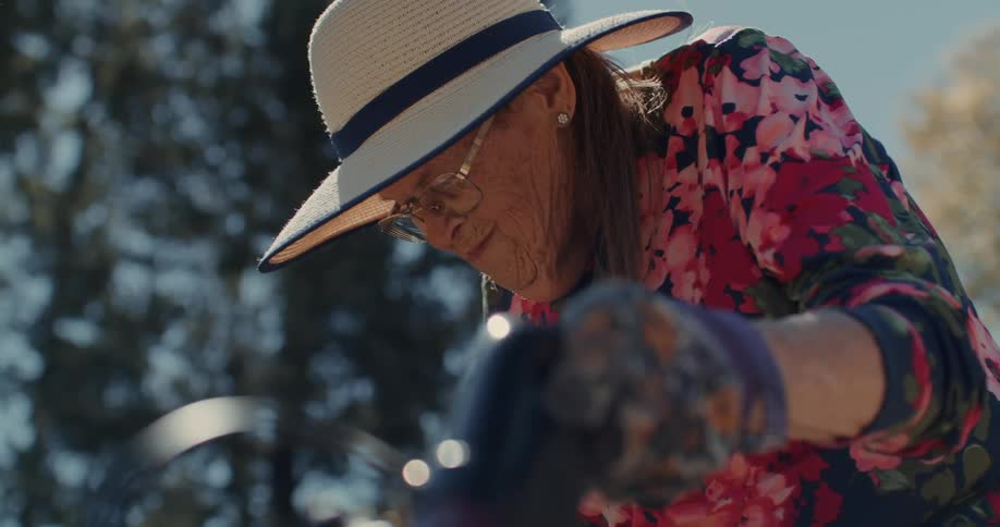 An elderly woman wearing a sun hat and gloves looking down while holding on to a handle