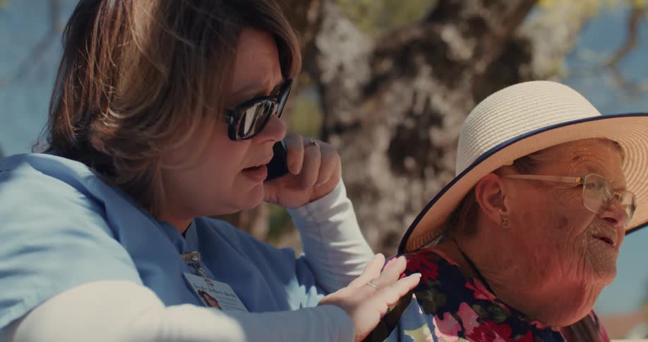 Female home caregiver pats elderly woman and talks on the phone while they sit outdoors