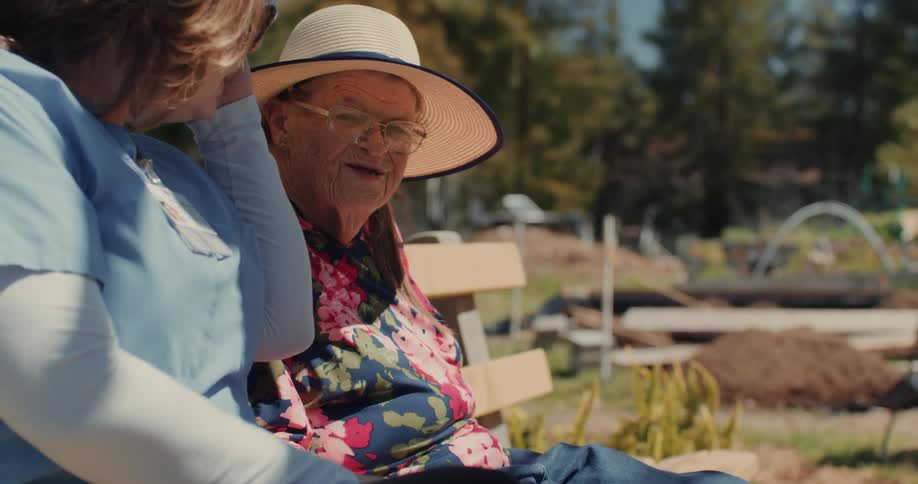 An elderly woman sits next to a nurse on a bench in the garden