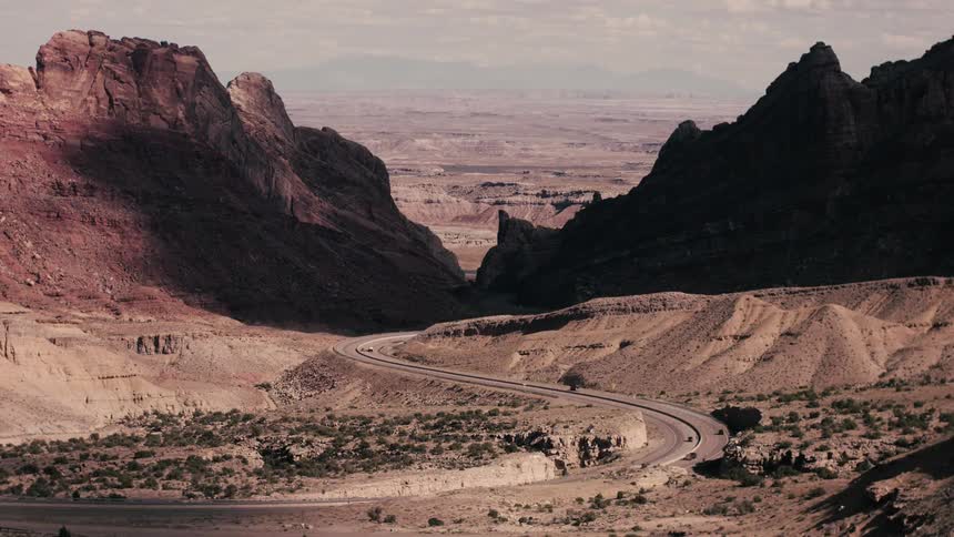 Traffic moves along highway winding through arid mountainous countryside while passing clouds cast shadows