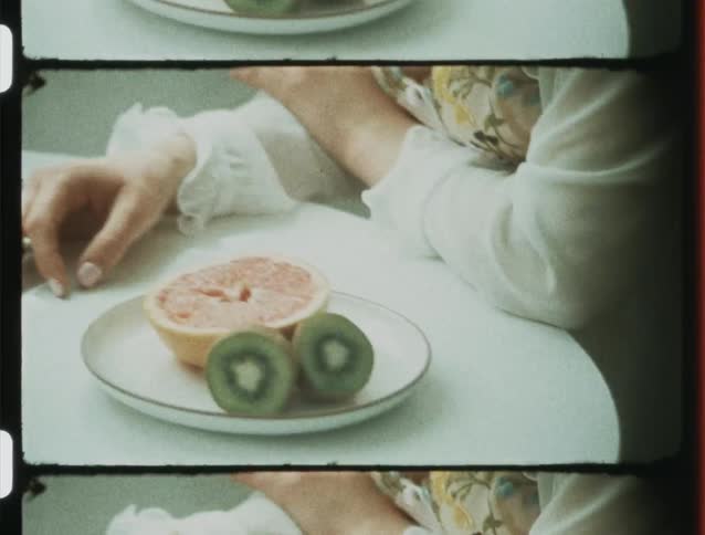 Hands of a woman sitting at a white table with a vase of flowers and a plate with sliced tangerines and kiwi