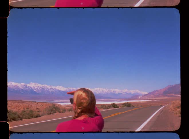 Blonde woman standing on highway shades her eyes while looking at natural scenery