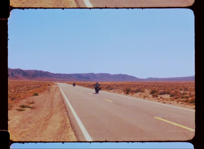Two motorbikes speeding past a paved road in a deserted location with mountains on the horizon