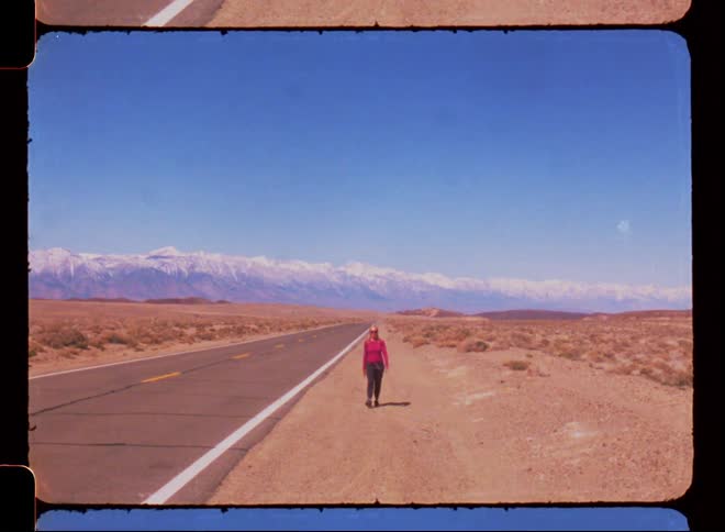 Woman walking along a paved road in a location with desert vegetation and mountains on the horizon