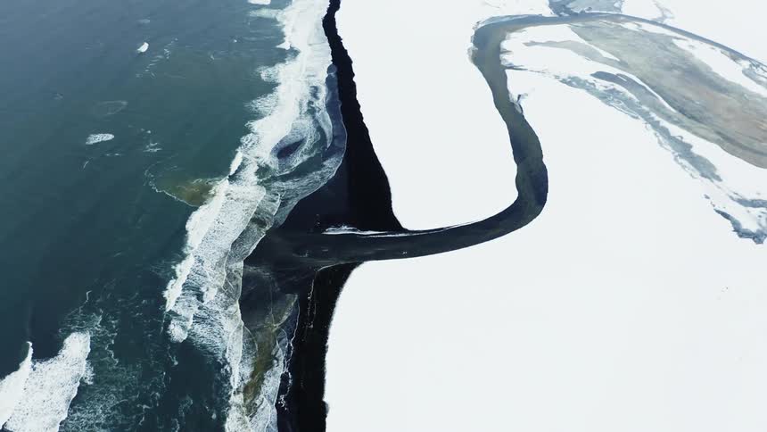 Dark river winds through snow covered coast as the waves roll onto the shore