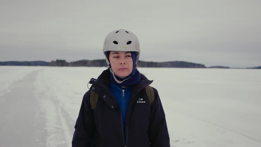 A young woman stands alone on an iced over lake in a snowy landscape wearing winter attire and a ski helmet