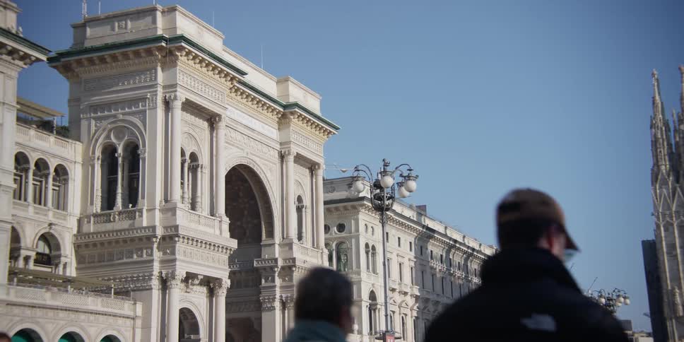 Pedestrians walk past a large building with a concrete archway on a sunny day