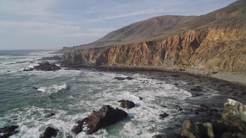 Vast sea with foamy waves crashing on the rocky coast of the mountains on a blue sky day with clouds