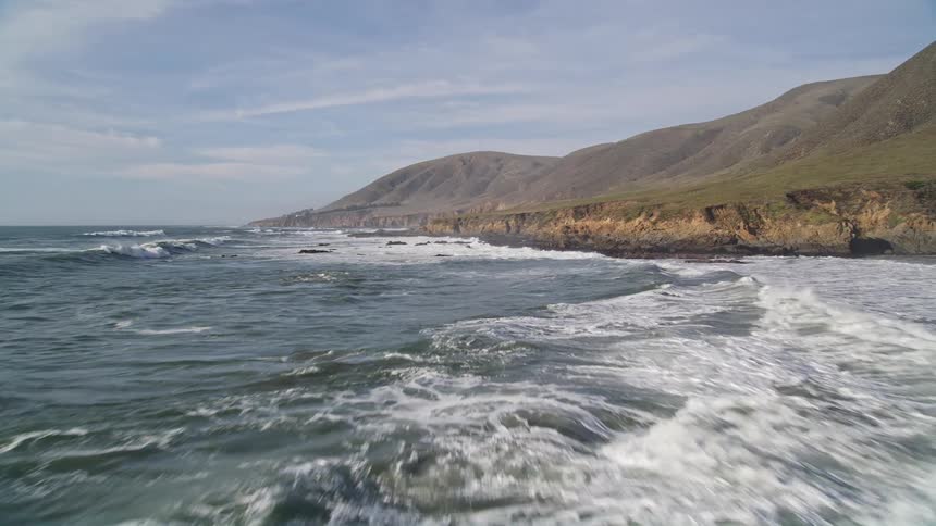 Rocky coastline lapped by ocean waves with mountains in the background under a clear sky