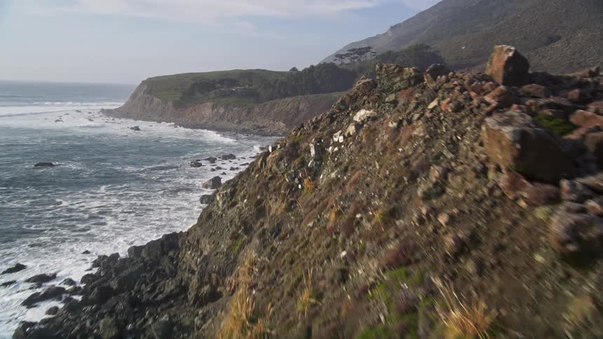 Sea waves crashing on the rocky coast with rural houses on hills during the day