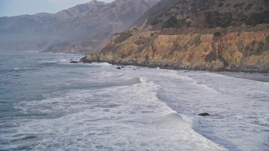 Cars drive down a coastal roads as the waves crash against the shore